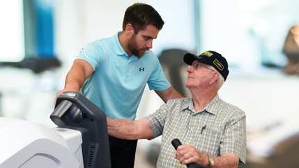 An older man in a black hat sitting at a machine while a McLeod Rehabilitation Specialist explains what he needs to do