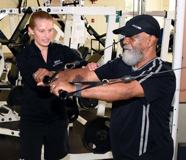 An older man using a chest press exercise machine at the McLeod Health & Fitness Center while a McLeod Health trainer supervises