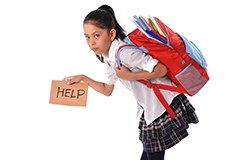 A young girl in a school uniform hunched over, carrying a backpack and holding a sign that says help
