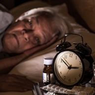 An older person with white hair laying awake in bed staring at a clock